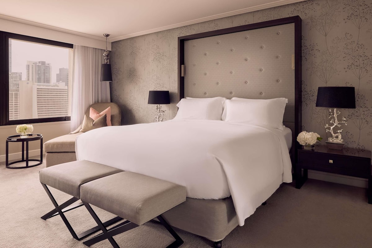 A hotel room with a large bed, two bedside tables, a chair, a bench, and a view of the city.
