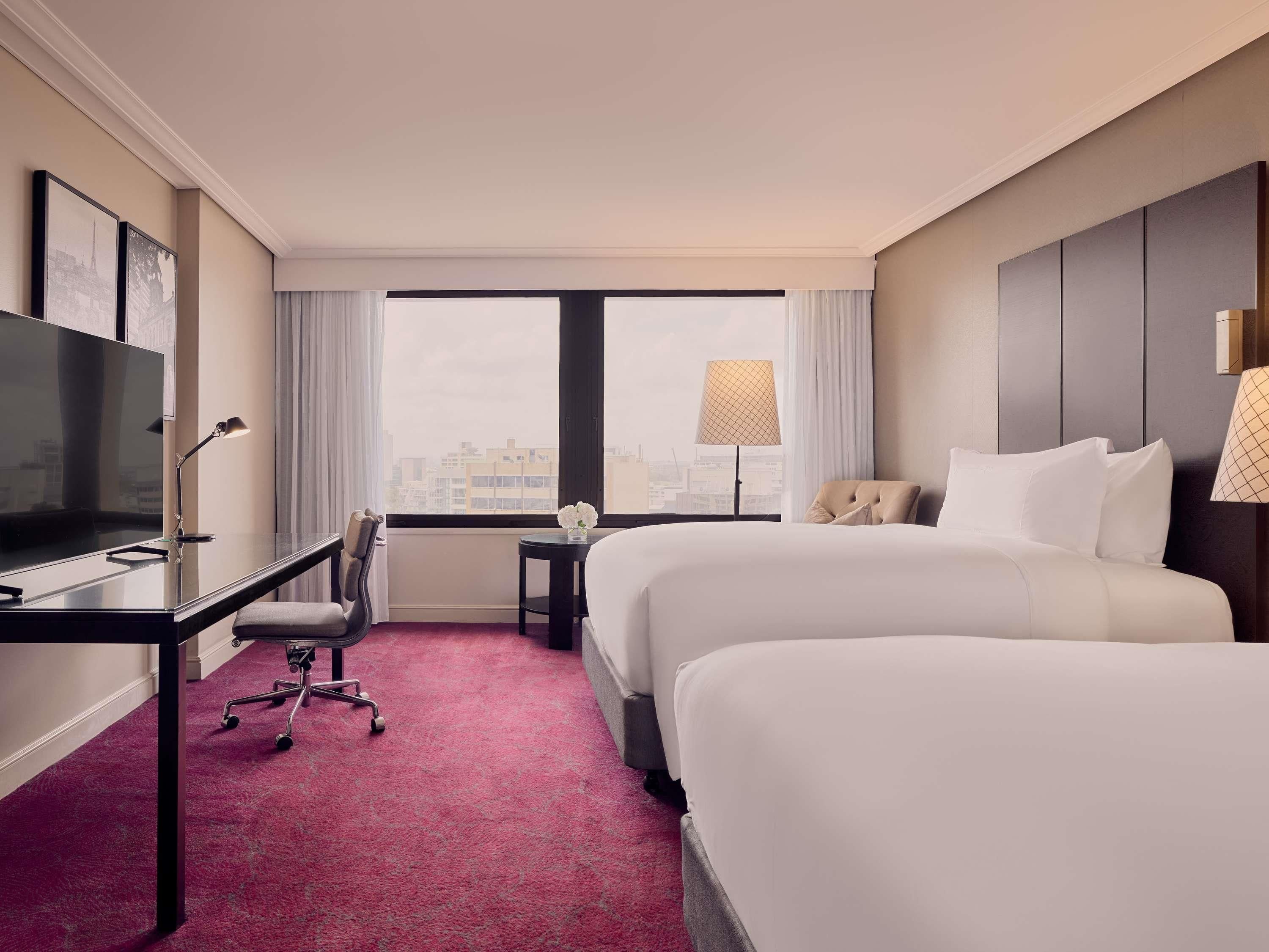 A hotel room with a bed, desk, chair, TV, lamp, and a view of the city.