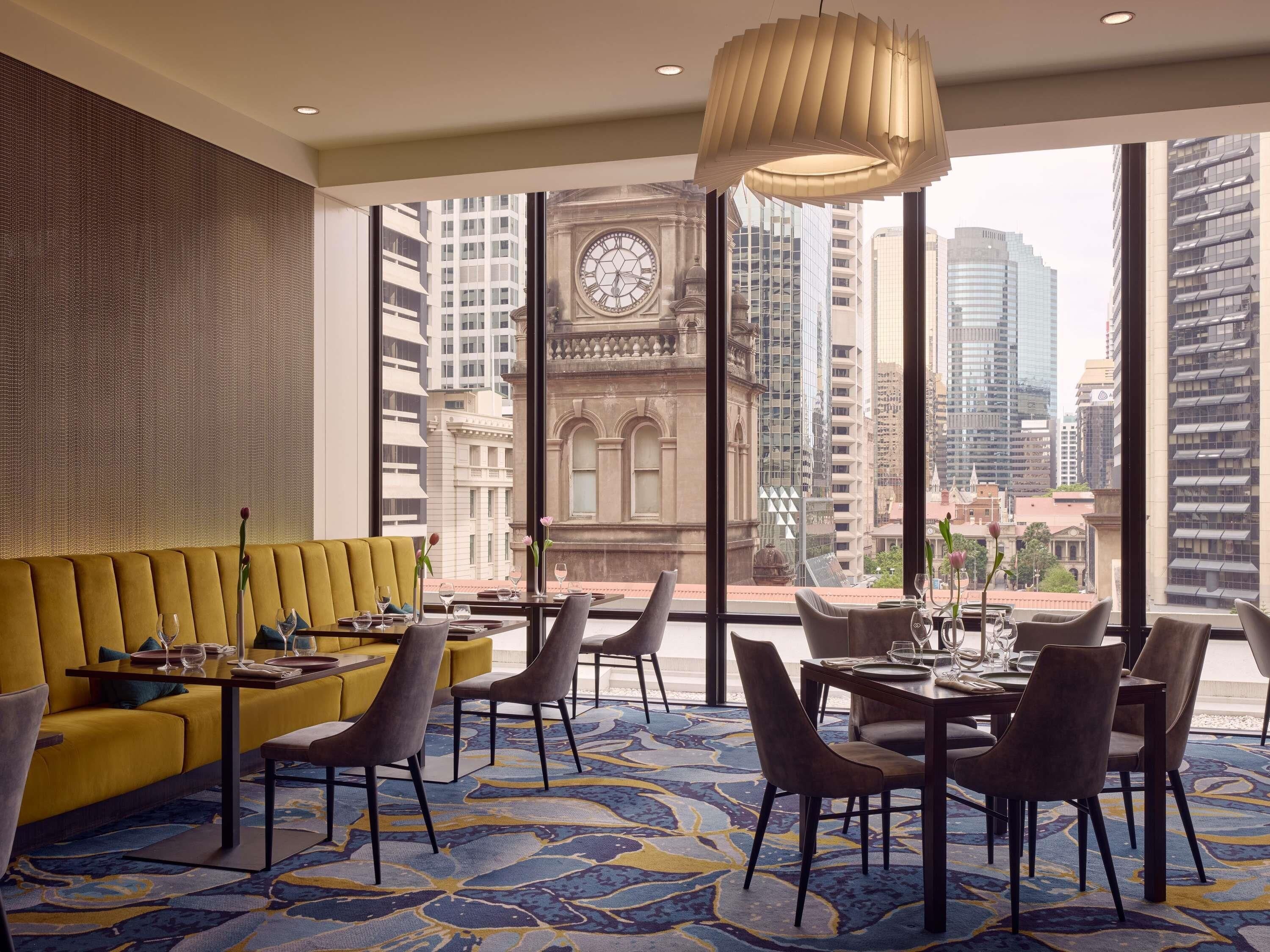 A modern hotel room with a yellow sofa, dining table, chairs, and a large window offering a city view.