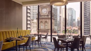 A modern hotel room with a yellow sofa, dining table, chairs, and a large window offering a city view.