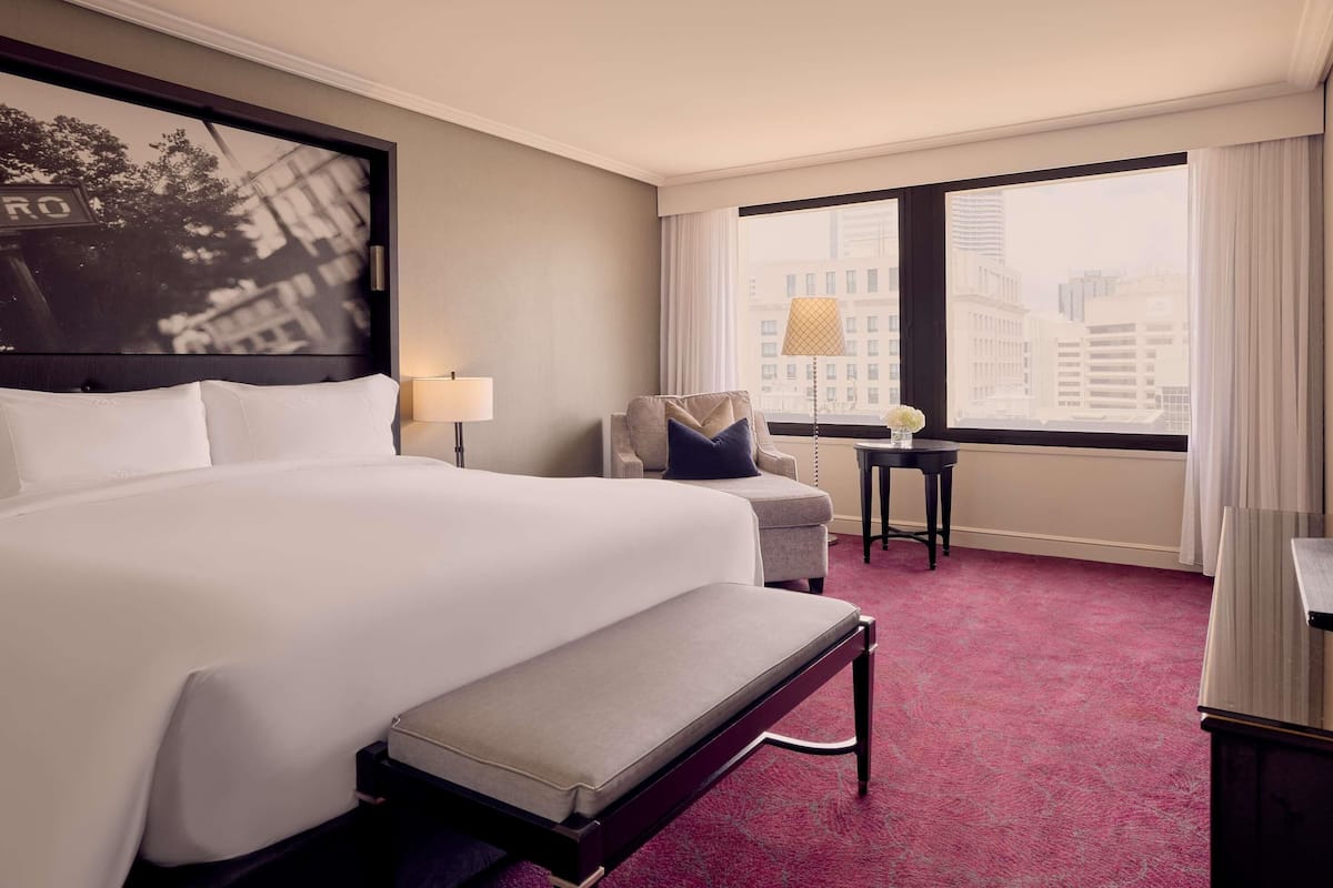 A hotel room with a large bed, a bench, a chair, a small table, and a view of the city through large windows.