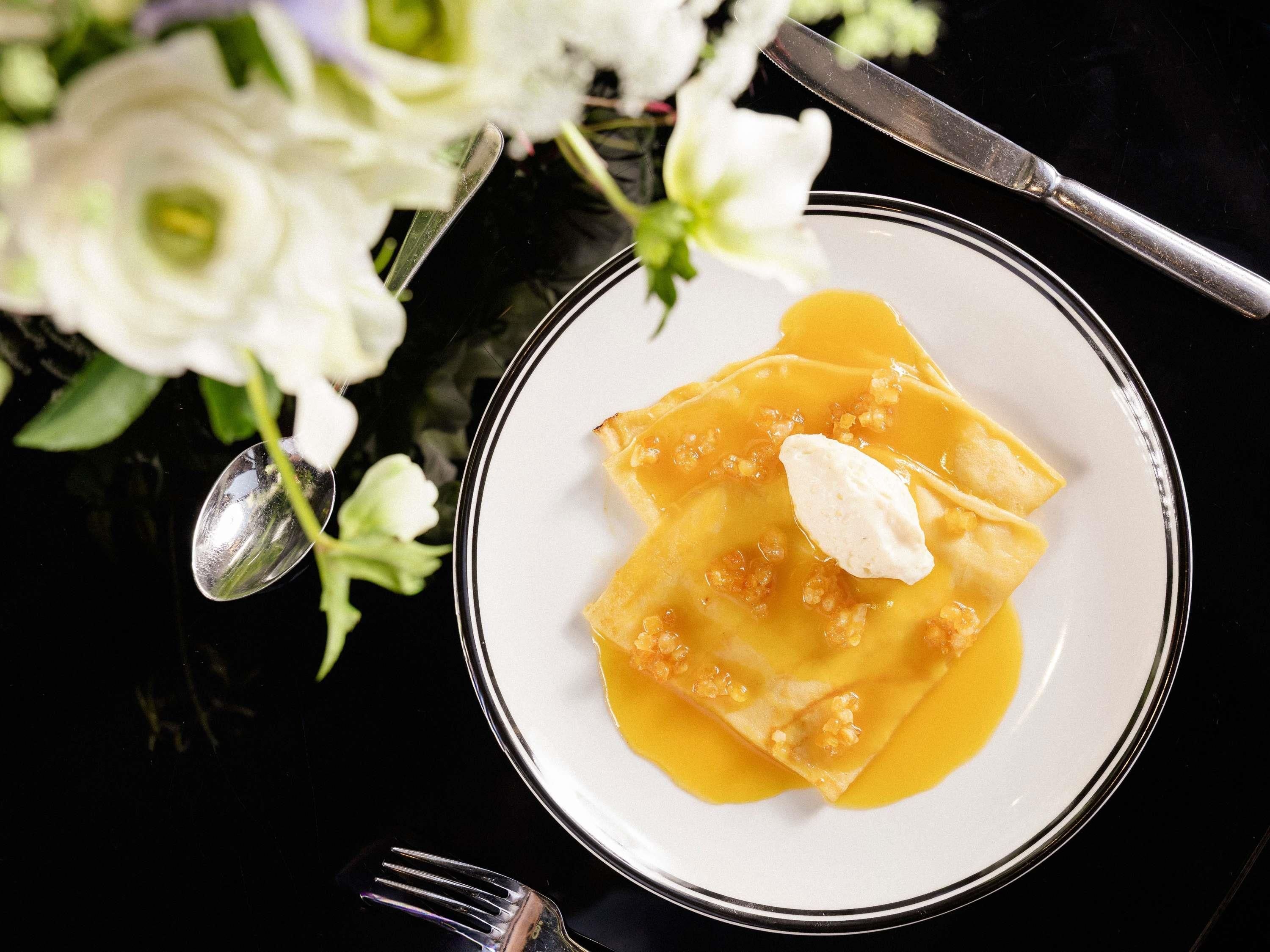 A plate of ravioli with sauce and a dollop of cream, accompanied by a fork and knife on a table with a black surface.