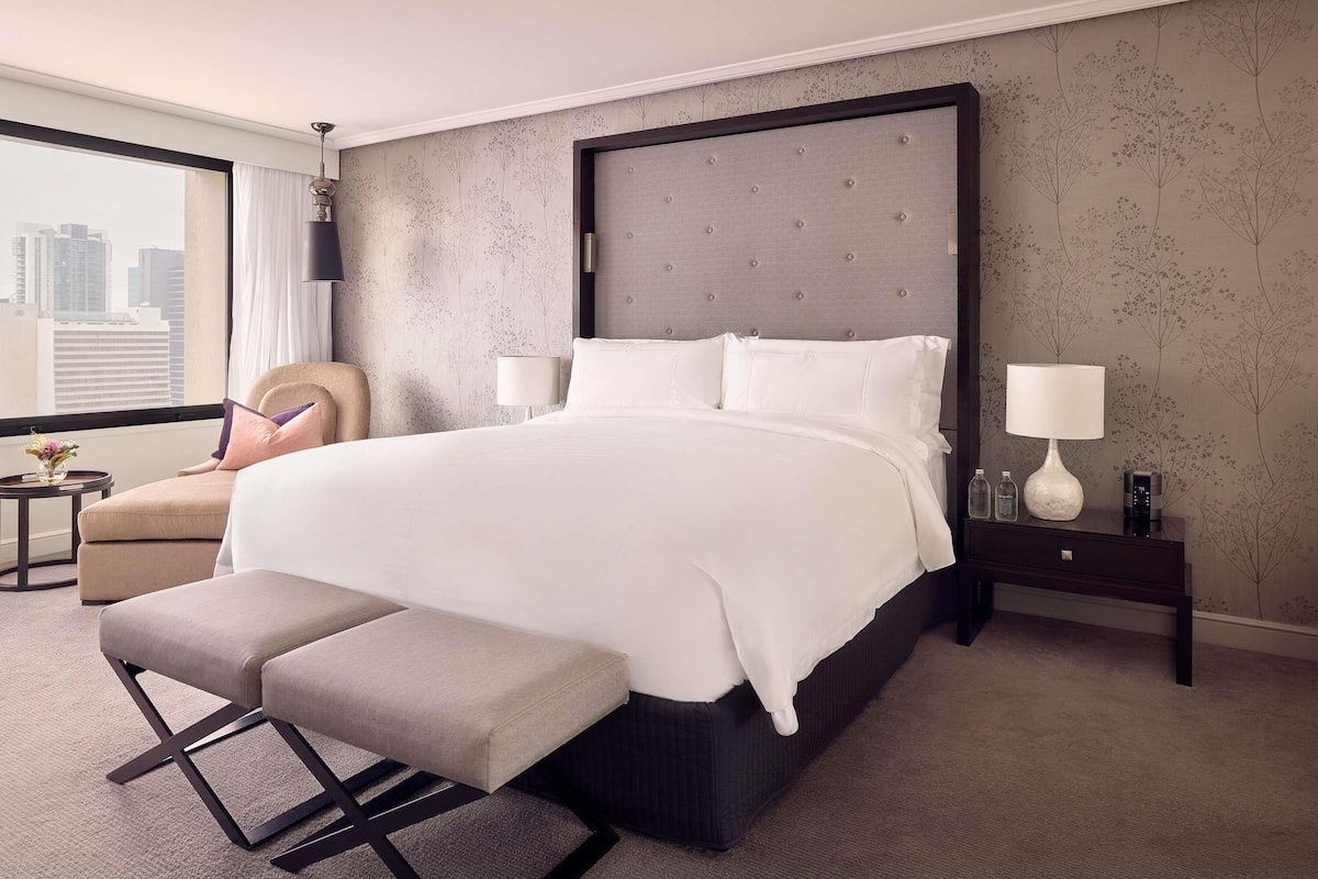 A hotel room with a large bed, a bench, a chair, a nightstand, and a view of the city.