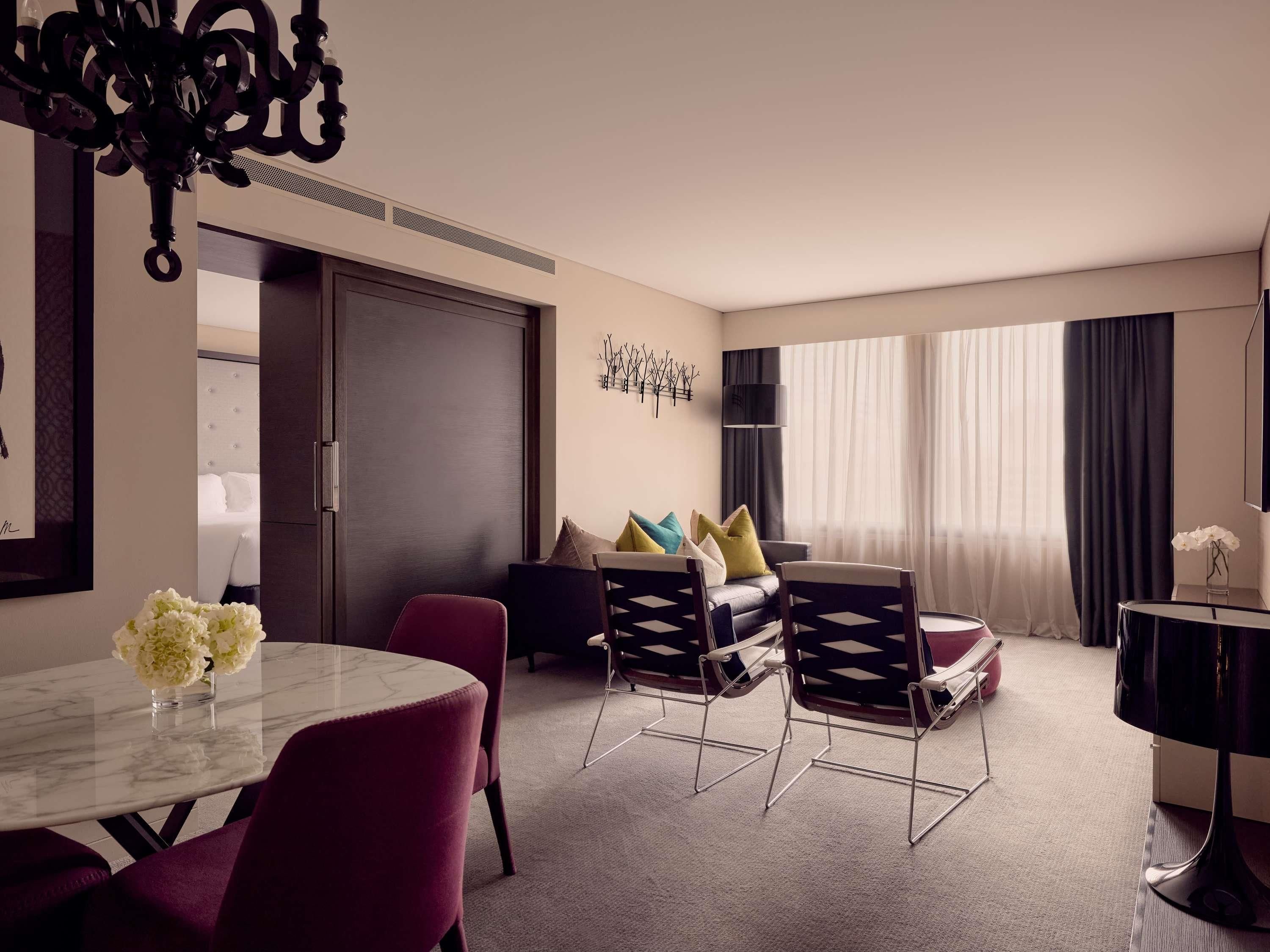 A hotel room with a dining area, a sofa set, a TV, and a large window with curtains.