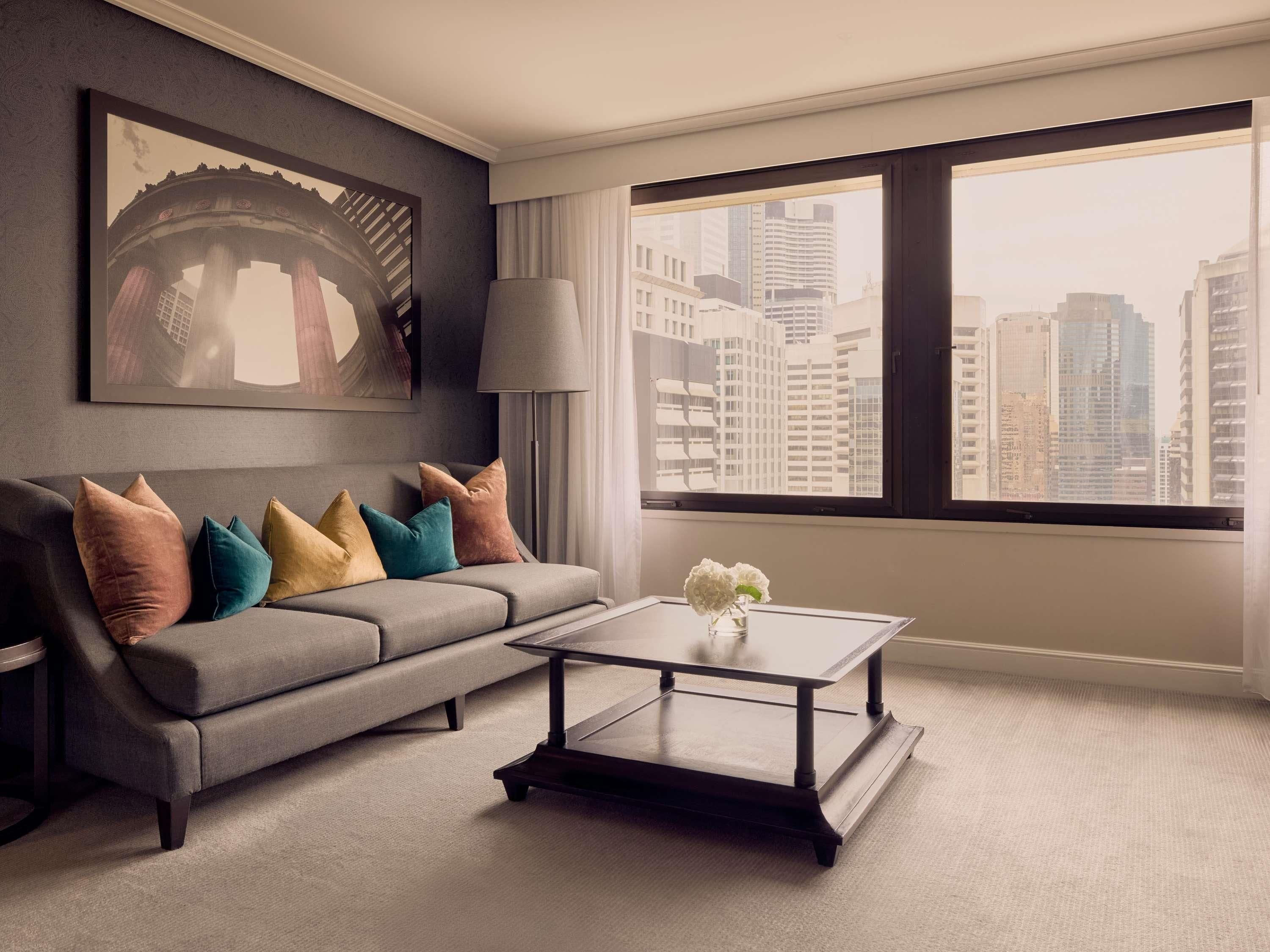 A modern living room with a grey sofa, colorful cushions, a glass coffee table, and a large window with a city view.