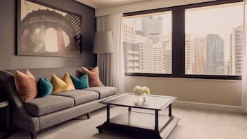 A modern living room with a grey sofa, colorful cushions, a glass coffee table, and a large window with a city view.