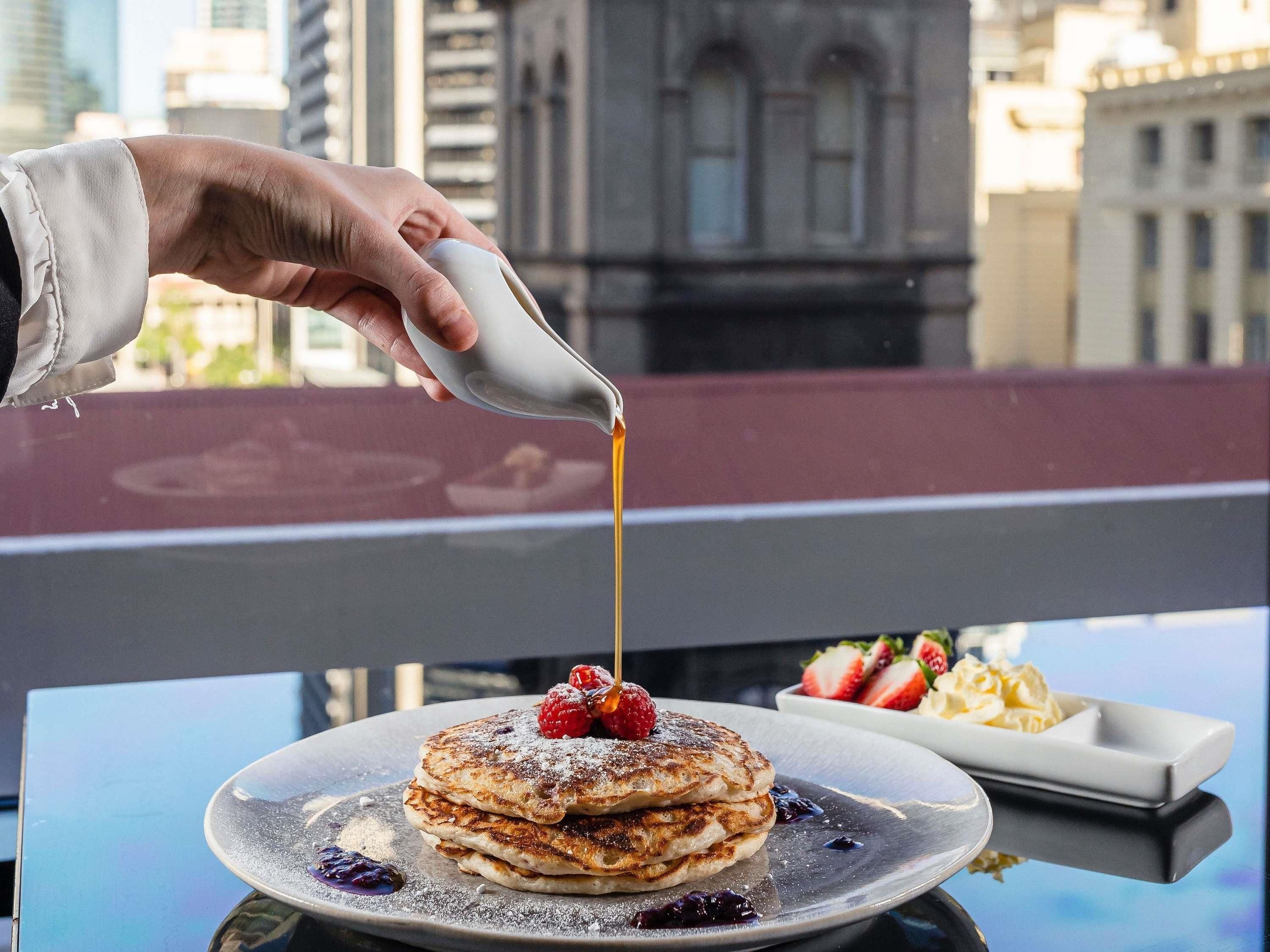 A hand pouring syrup onto pancakes on a plate, with a dessert tray and a cityscape in the background.