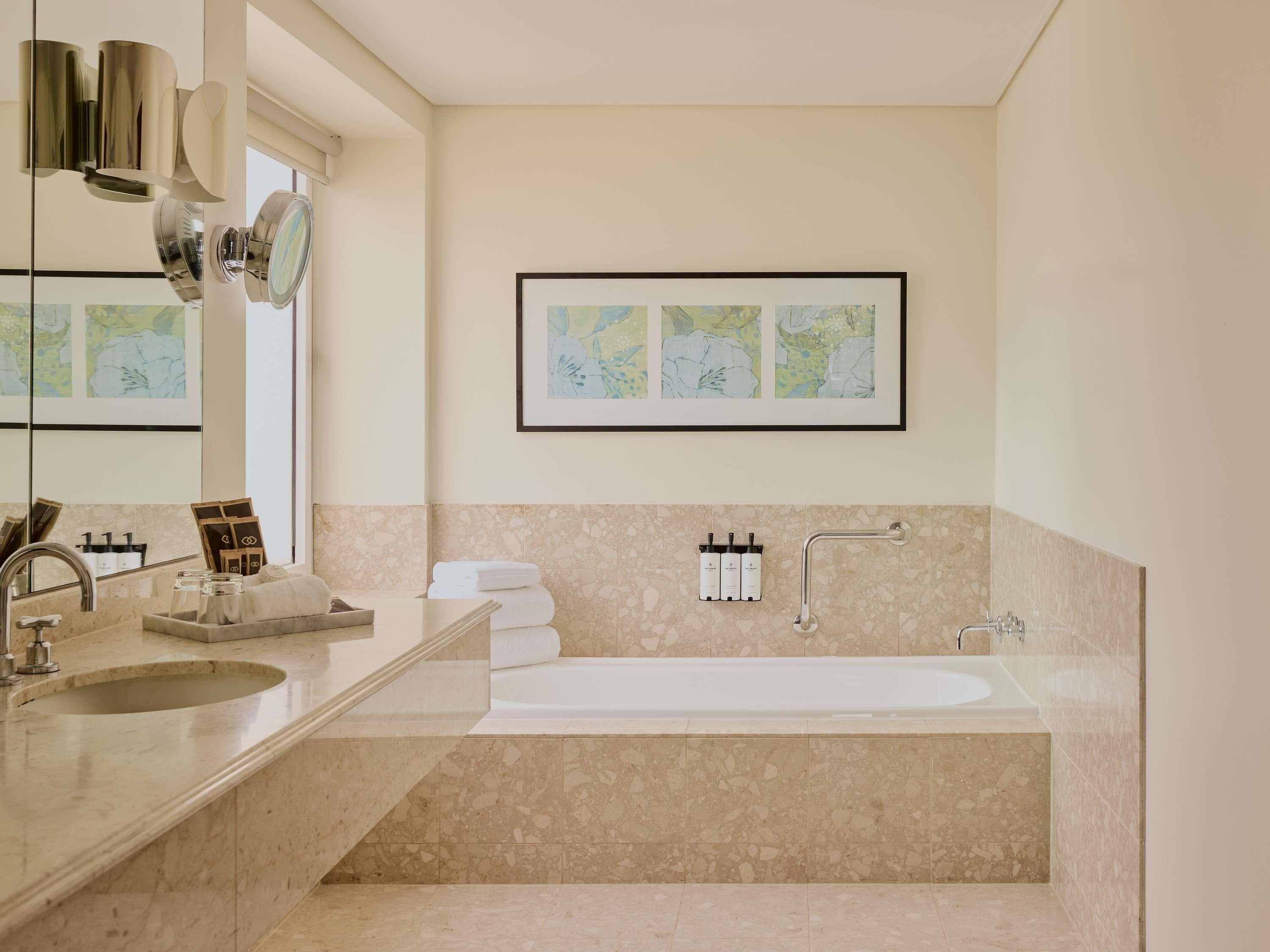 Une salle de bain moderne avec une grande baignoire, un lavabo et un miroir. Des serviettes sont pliées et empilées sur un porte-serviettes mural, et un tableau encadré est accroché au-dessus de la baignoire.