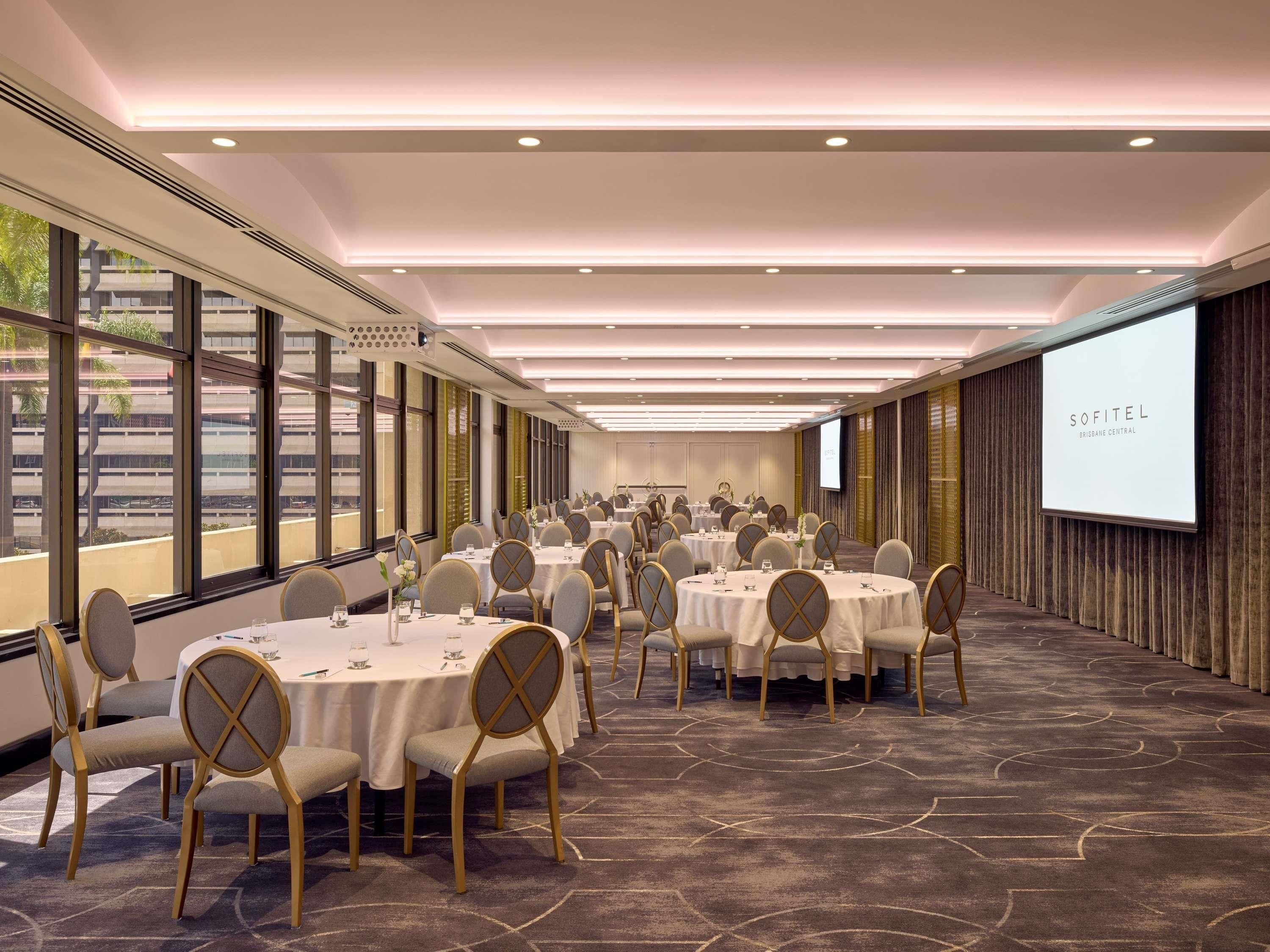 Un salón de conferencias con mesas redondas, sillas y una gran pantalla mostrando 'SOFITEL BRISBANE CENTRAL'.