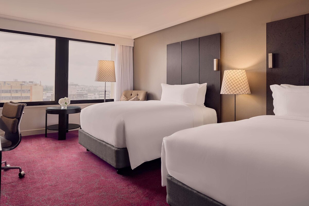 A hotel room with two beds, a desk, a chair, and a view of the city.