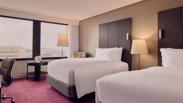A hotel room with two beds, a desk, a chair, and a view of the city.