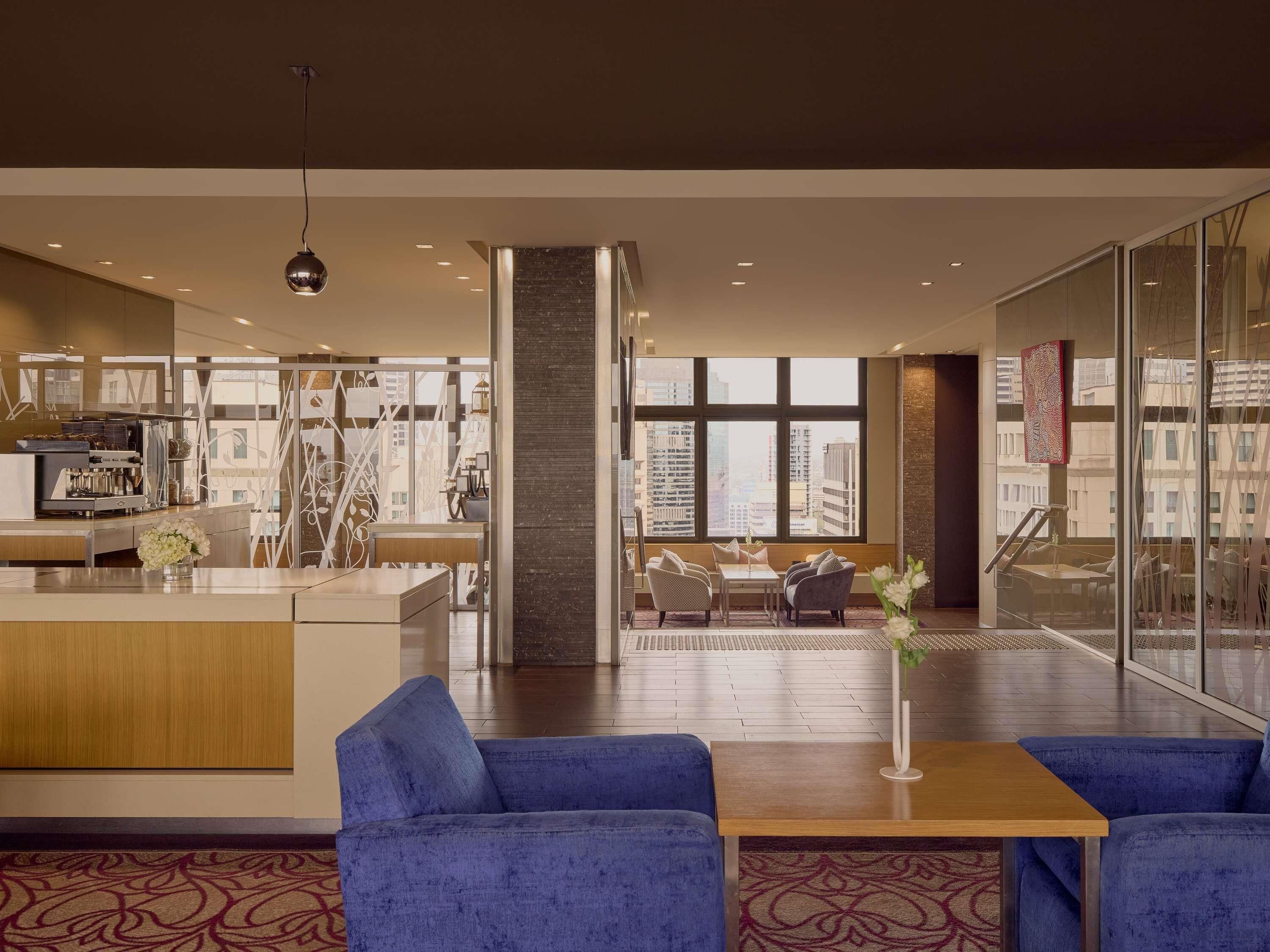 A modern hotel lobby with a central bar, seating area, and large windows offering city views.