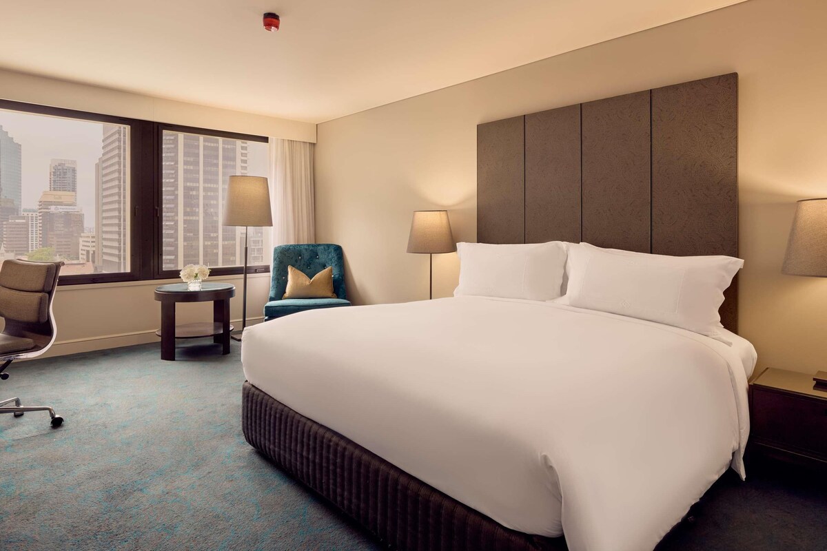 A hotel room with a large bed, two bedside lamps, a chair, a small table, and a view of a cityscape through large windows.