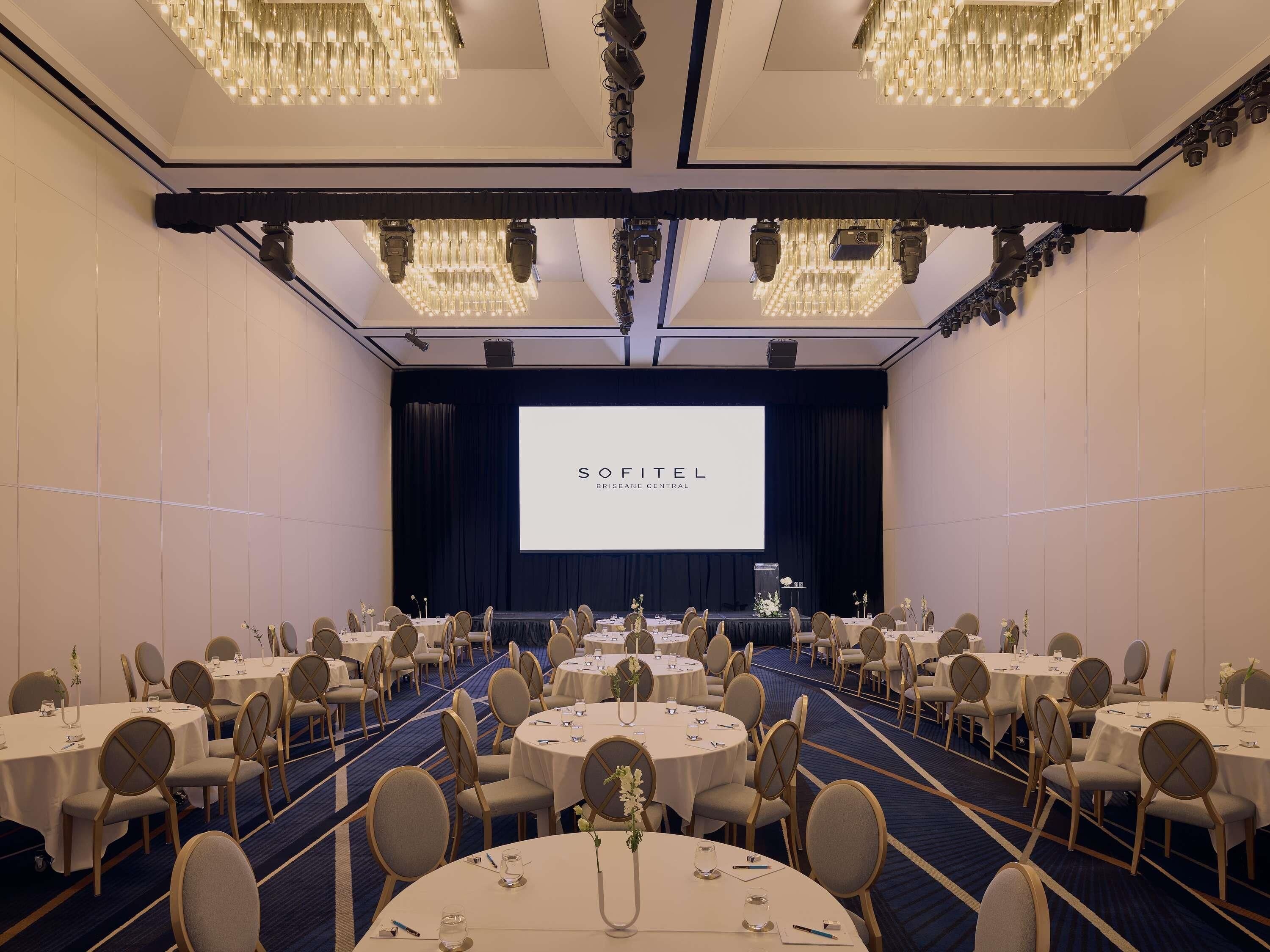 A conference room with round tables, chairs, and a large screen displaying 'SOFITEL BRISBANE CENTRAL'.