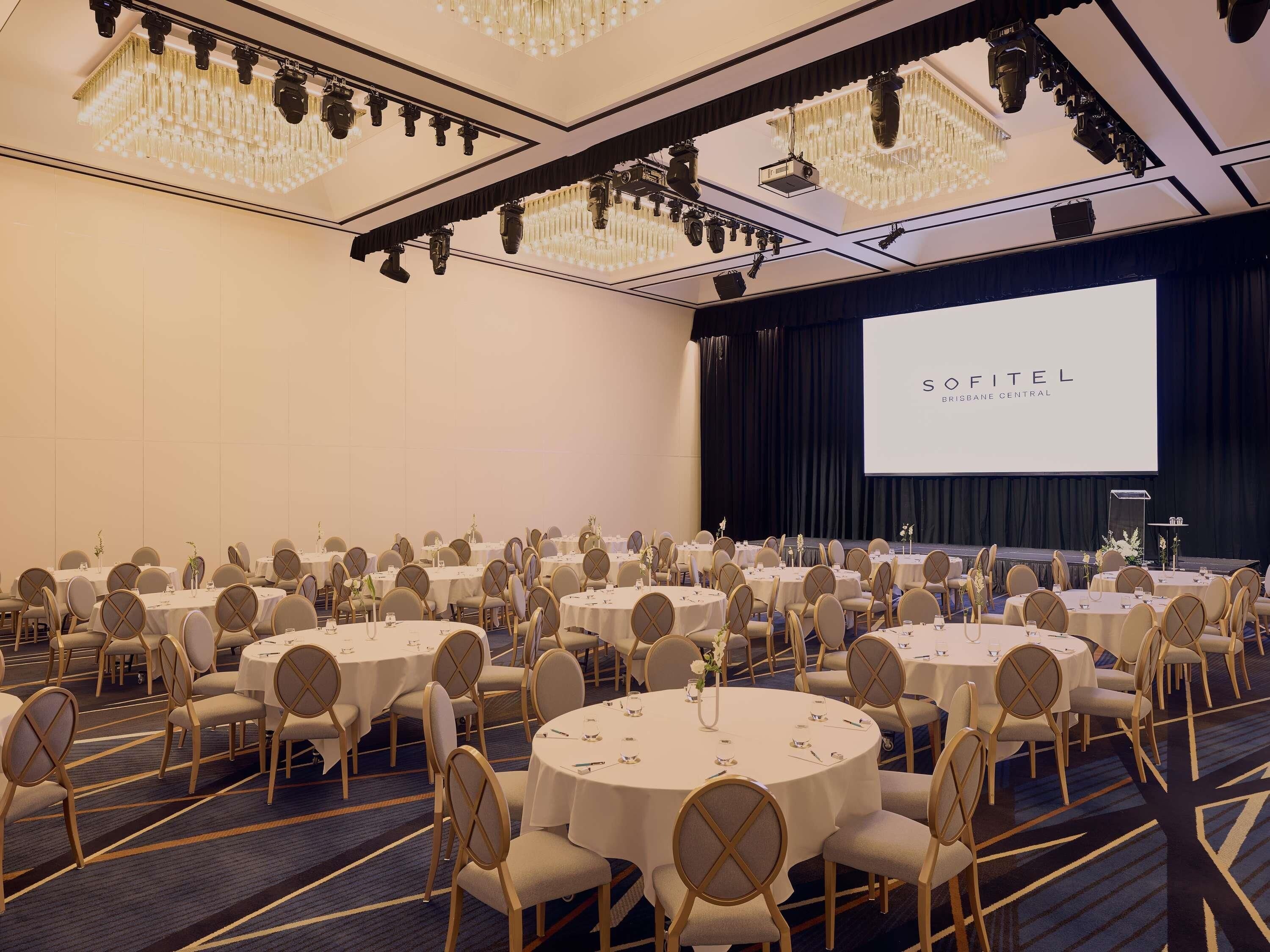 Een vergaderzaal met ronde tafels, stoelen, een podium en een groot scherm.