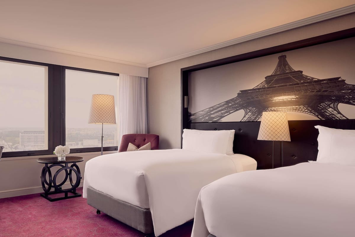 A hotel room with two beds, a view of the Eiffel Tower, a red chair, and a small table with a lamp.