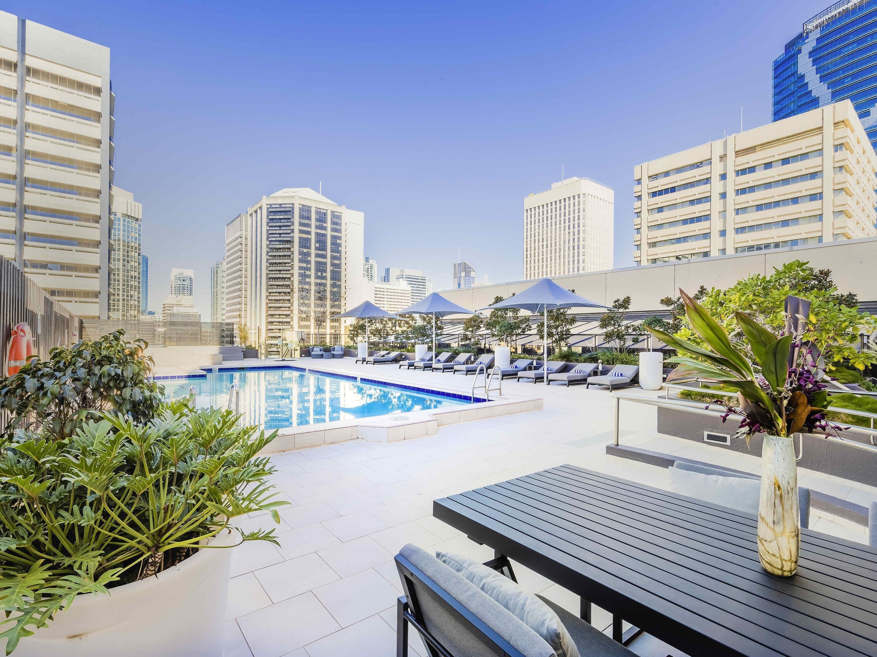 A rooftop pool area with lounge chairs, a pool, and city buildings in the background.