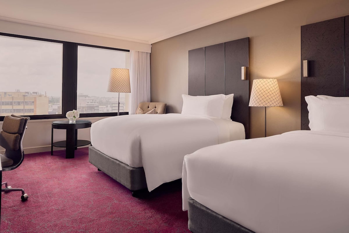 A hotel room with two beds, a desk, a chair, and a view of the city.