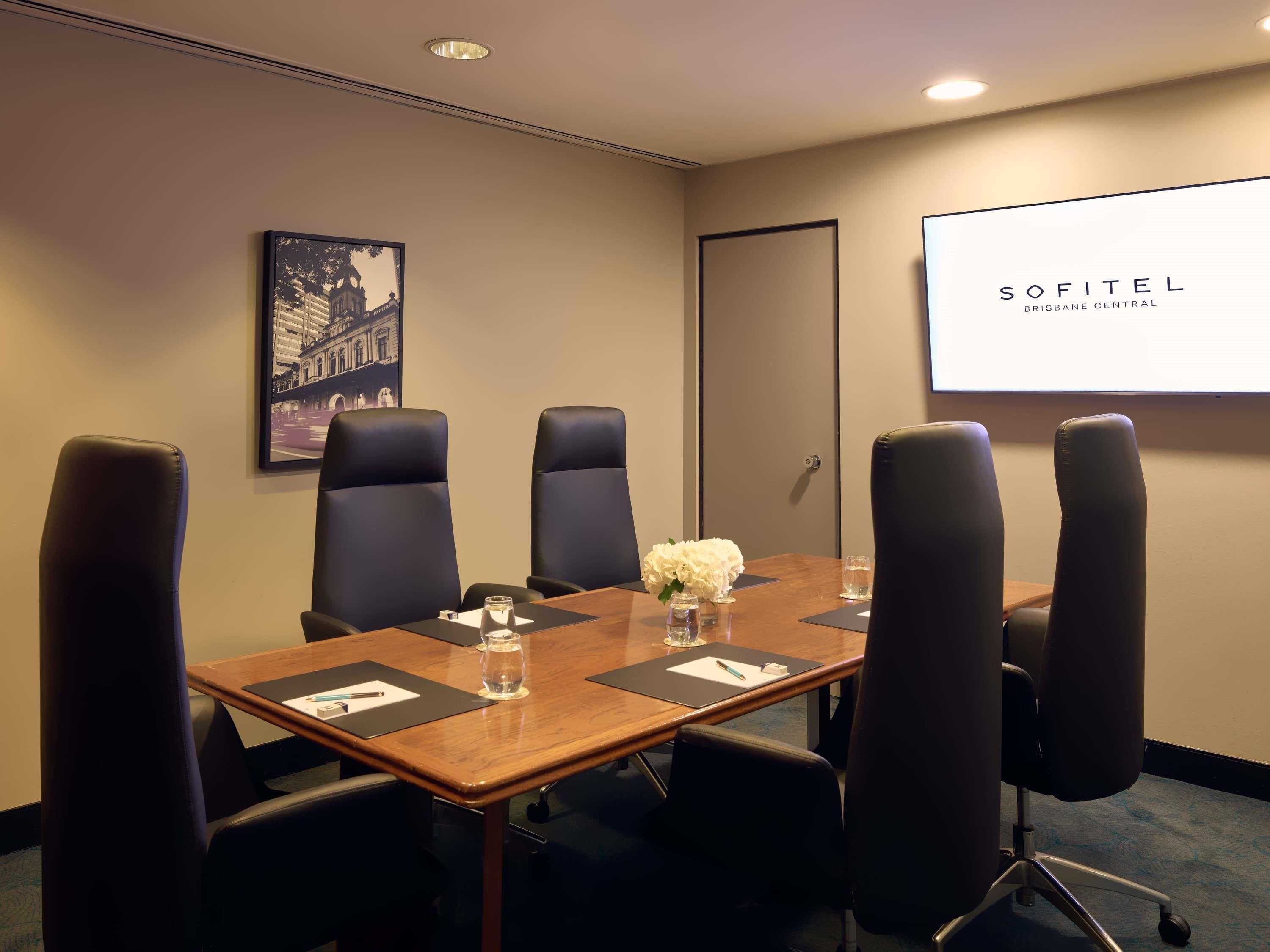 A conference room with a wooden table, six black chairs, a projector screen displaying 'SOFITEL BRISBANE CENTRAL', and a framed picture on the wall.