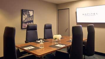 A conference room with a wooden table, six black chairs, a projector screen displaying 'SOFITEL BRISBANE CENTRAL', and a framed picture on the wall.