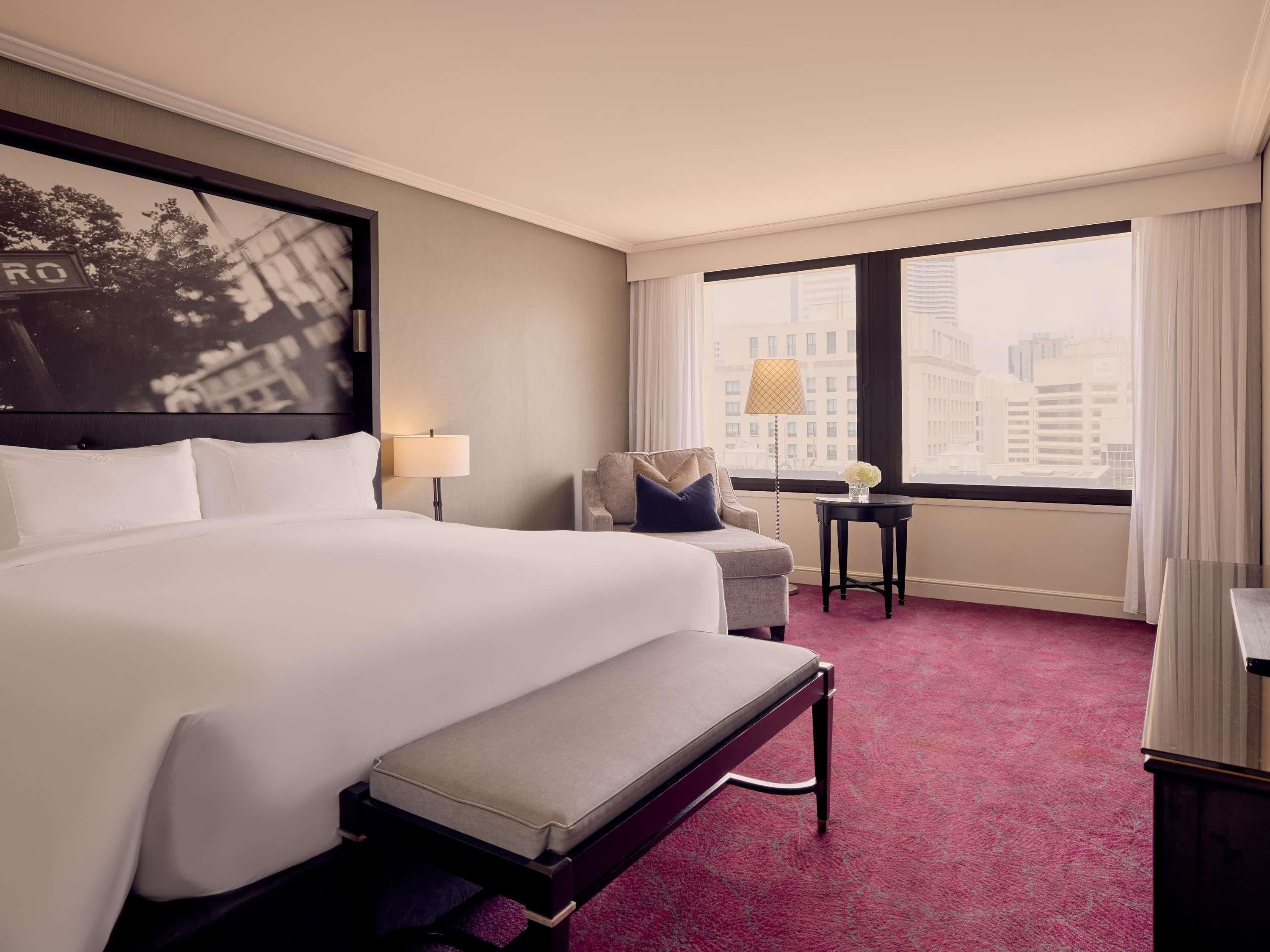 A hotel room with a large bed, a bench, a chair, a small table, and a view of the city through large windows.