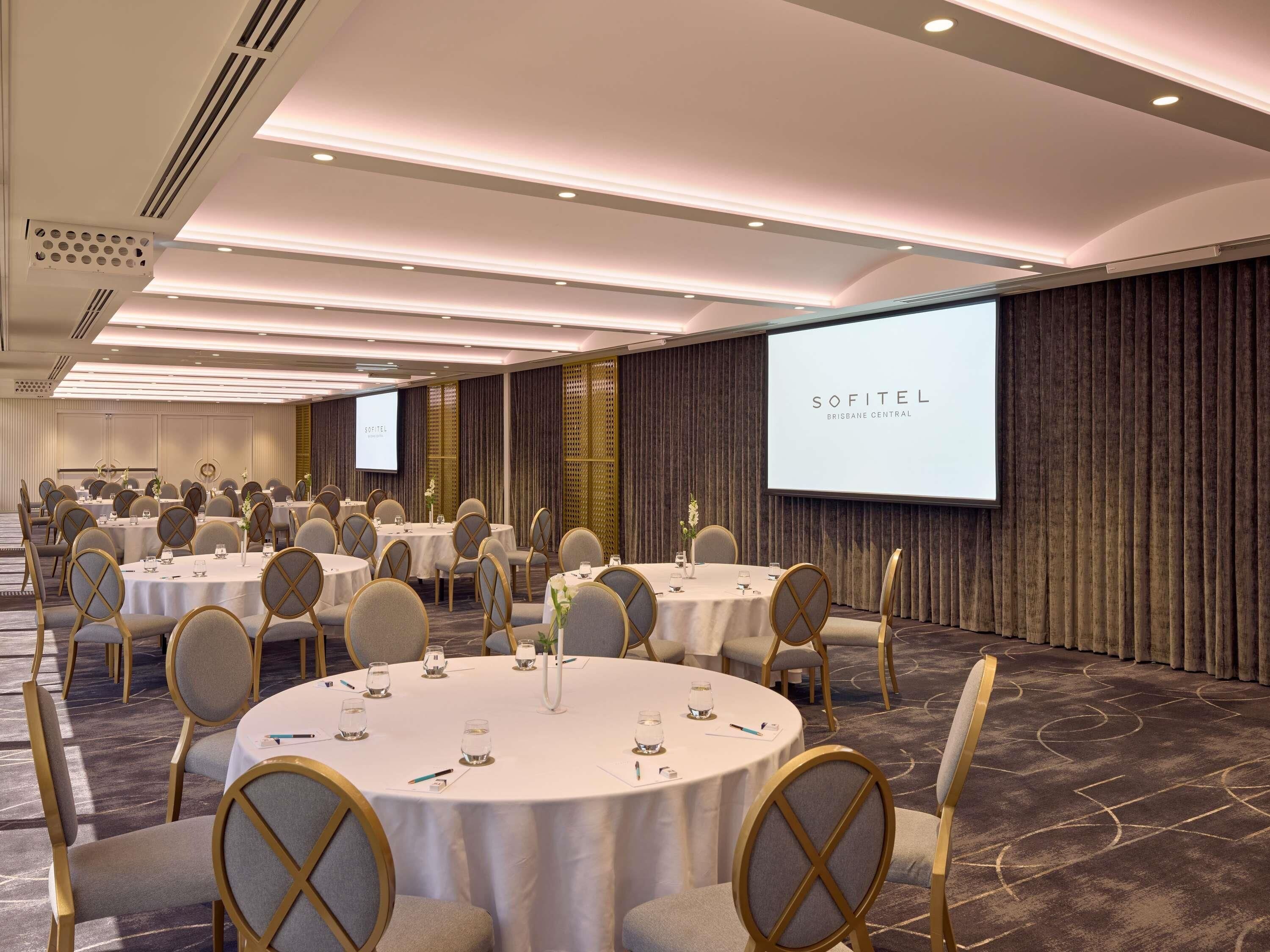 A conference room with round tables, chairs, and a projection screen displaying 'SOFITEL BRISBANE CENTRAL'.