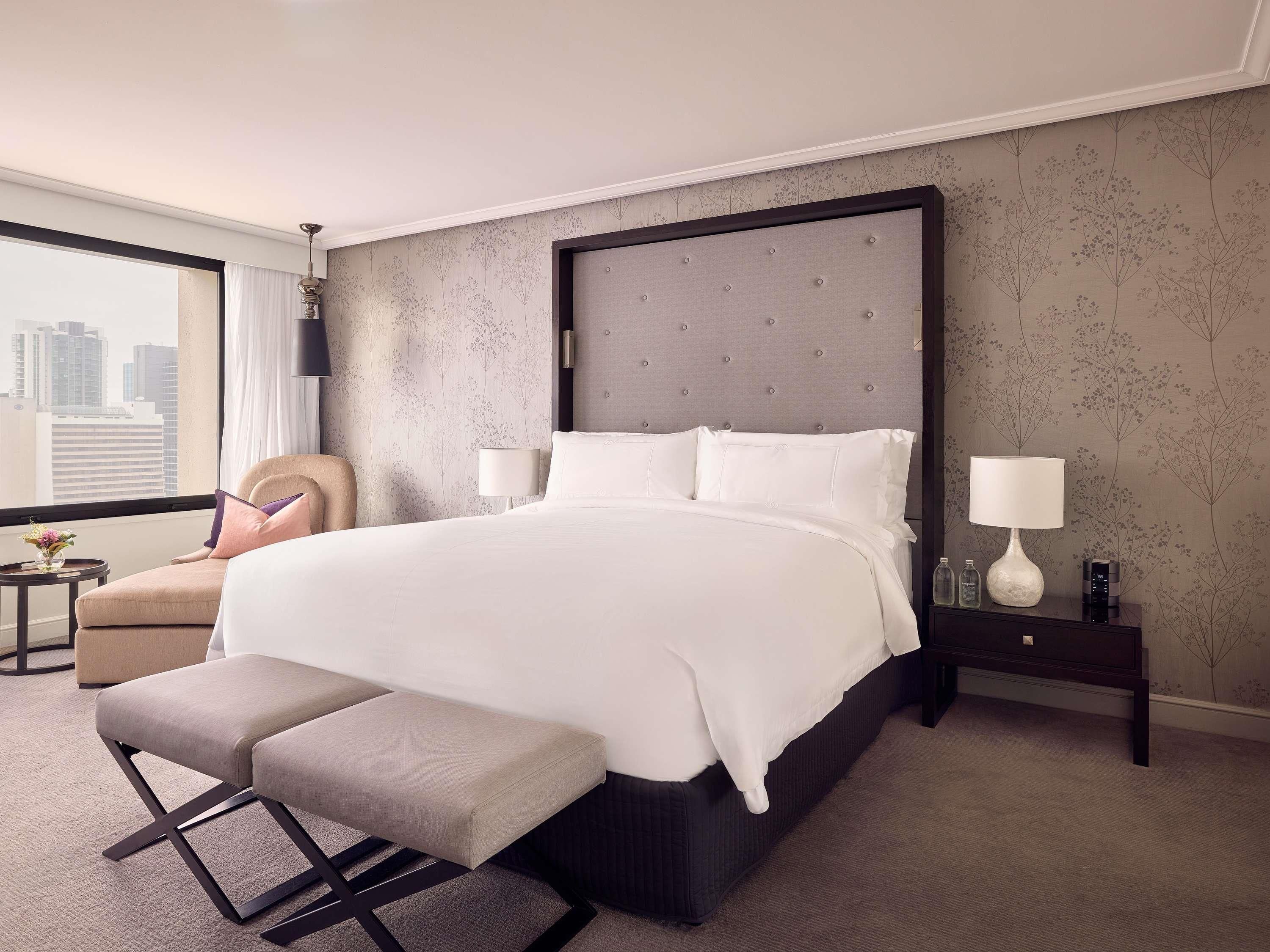 A hotel room with a large bed, a bench, a chair, a nightstand, and a view of the city.
