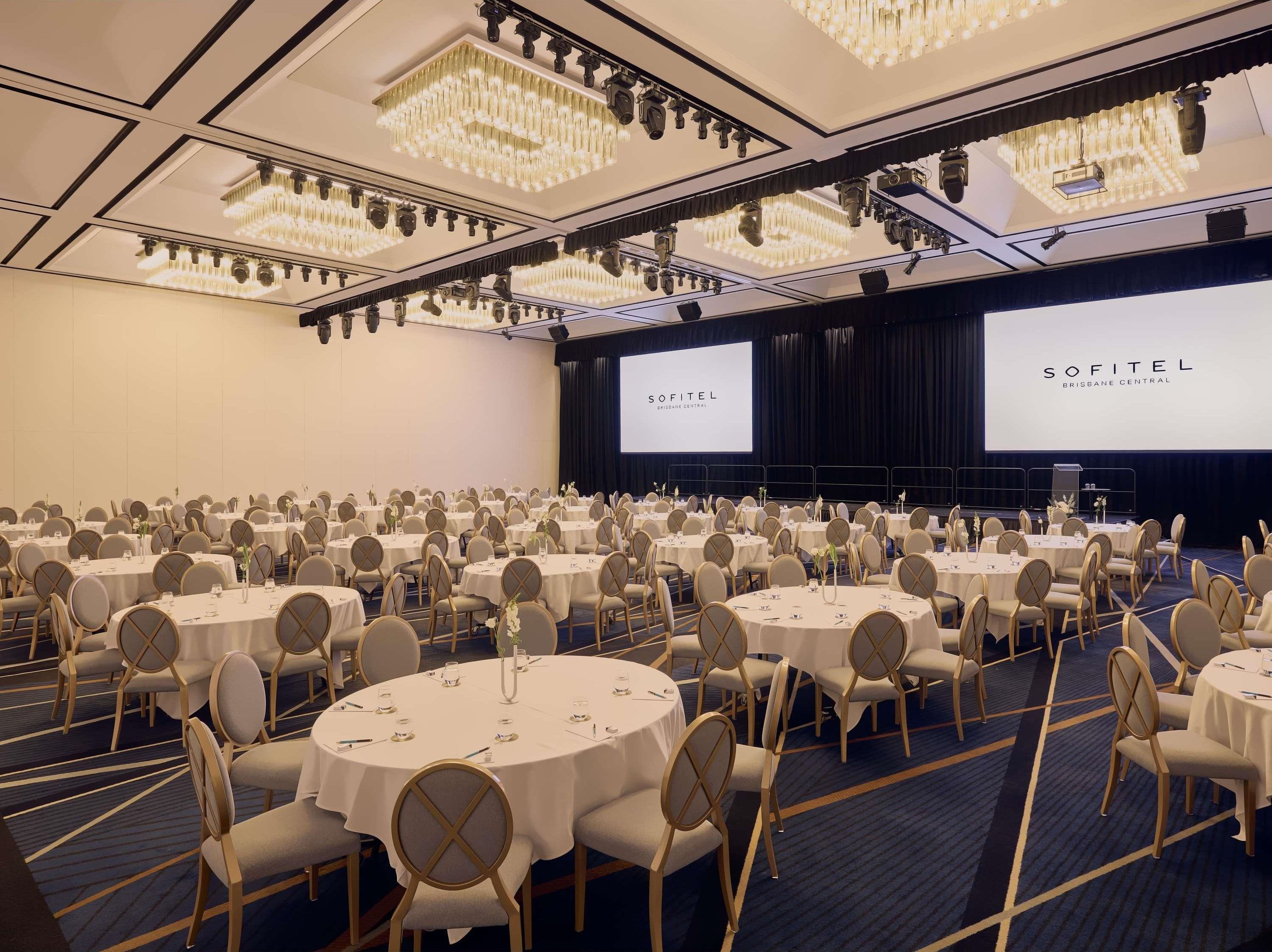 Kokoustila Sofitel Brisbane Central -hotellissa, jossa on pyöreitä pöytiä, tuoleja ja koroke, jossa on projektorikangas.