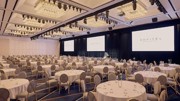 A large conference room with round tables, chairs, and a stage with screens displaying 'SOFITEL BRISBANE CENTRAL'.