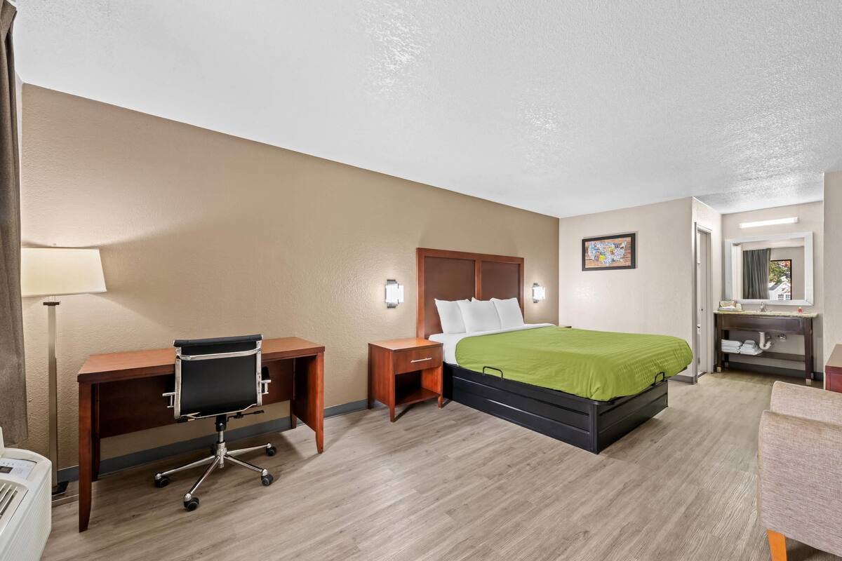 A hotel room with a bed, desk, chair, bedside table, lamp, and a bathroom with a sink and mirror.
