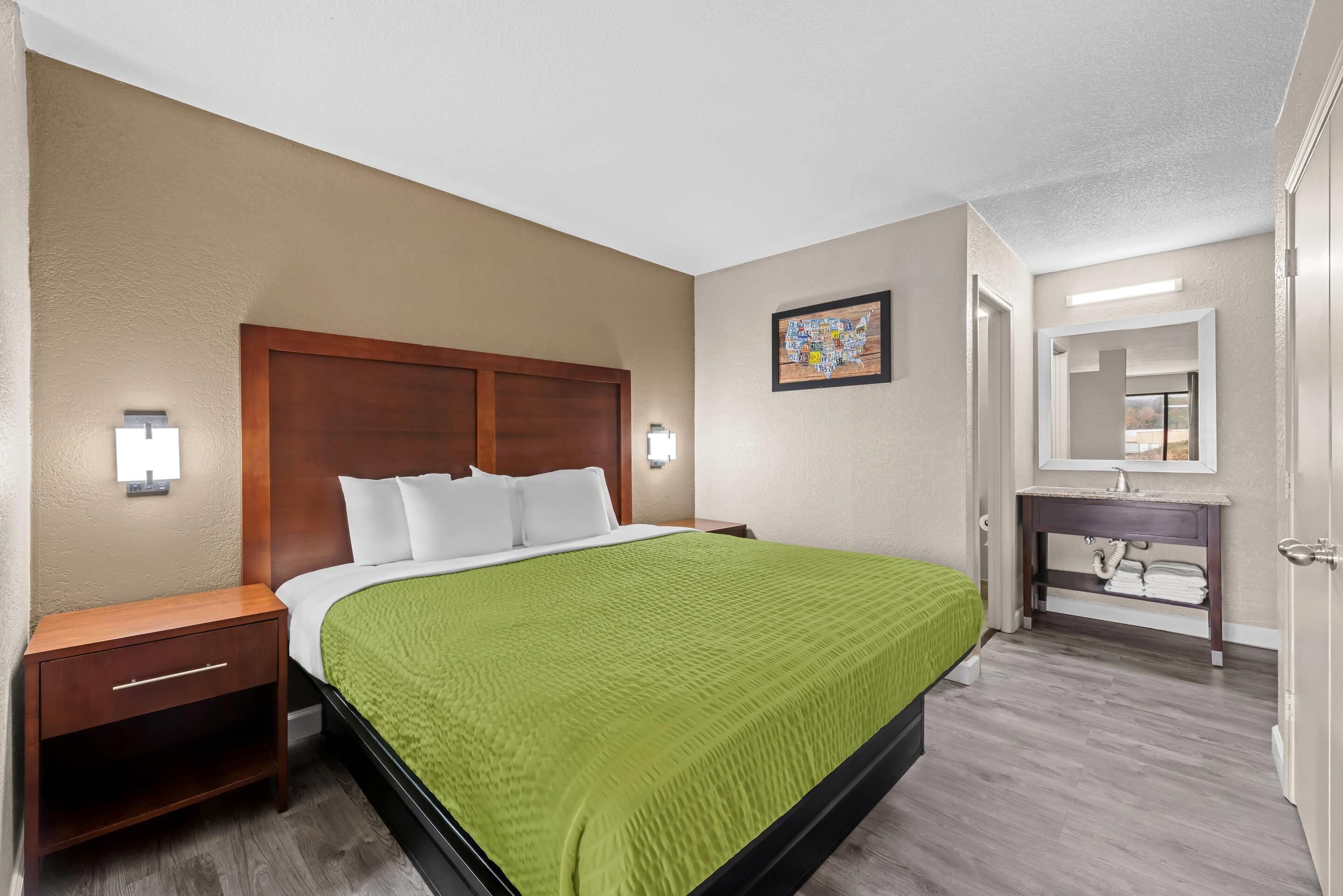 Habitación de hotel con cama, mesita de noche, tocador y lavamanos en el baño.
