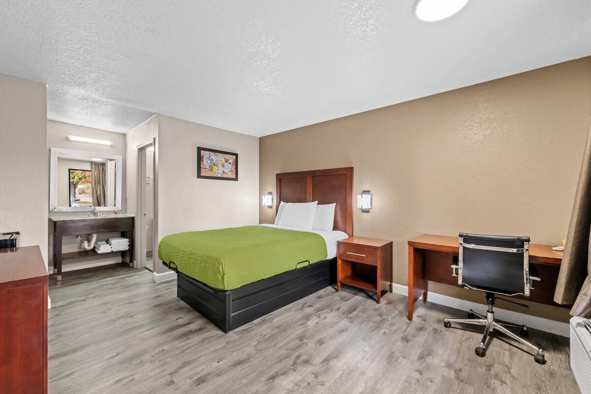A hotel room with a bed, bedside table, desk, chair, and a bathroom with a sink and mirror.