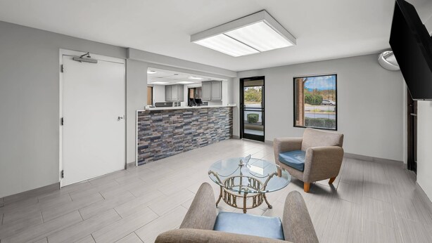 A modern living room with a glass coffee table, two armchairs, a kitchen area with a stone backsplash, and a large window offering a view of the outdoors. - Econo Lodge Kannapolis - Concord (Kannapolis)
