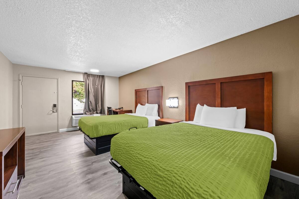 A hotel room with two beds, a desk, a window with curtains, and a door.