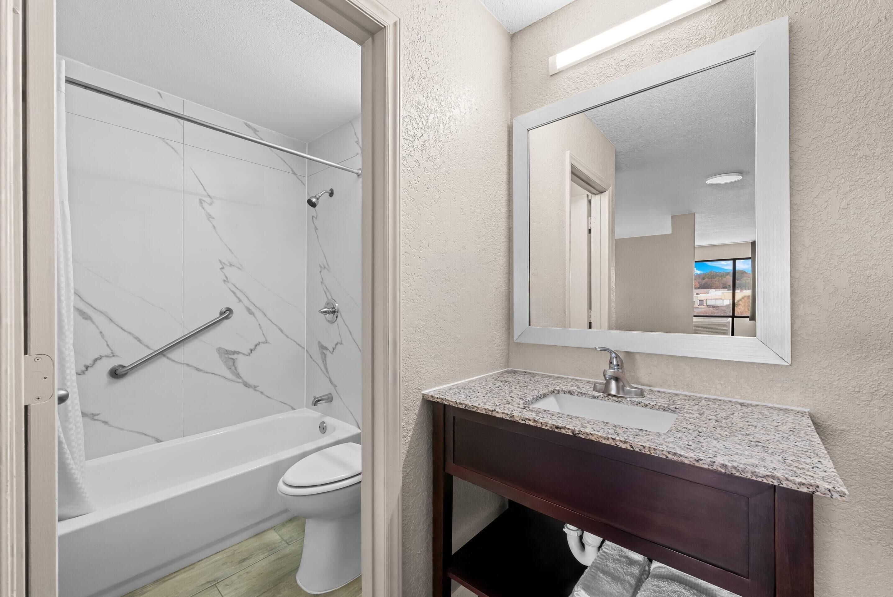 Un baño moderno con pared de ducha de mármol, inodoro blanco, tocador de madera oscura y un espejo grande.