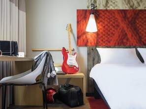 A hotel room with a bed, a desk with a guitar, a chair, a suitcase, and a red helmet. - ibis Hotel Köln Am Dom (Cologne)