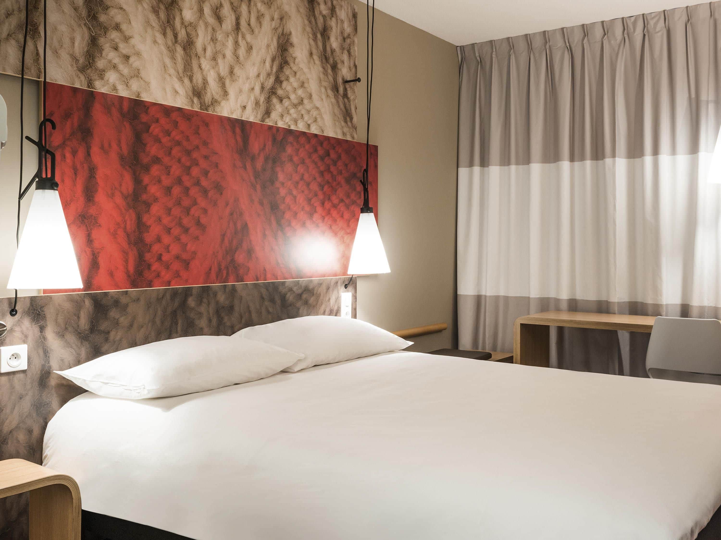 A hotel room with a bed, two bedside lamps, a desk, a chair, and a window with curtains.