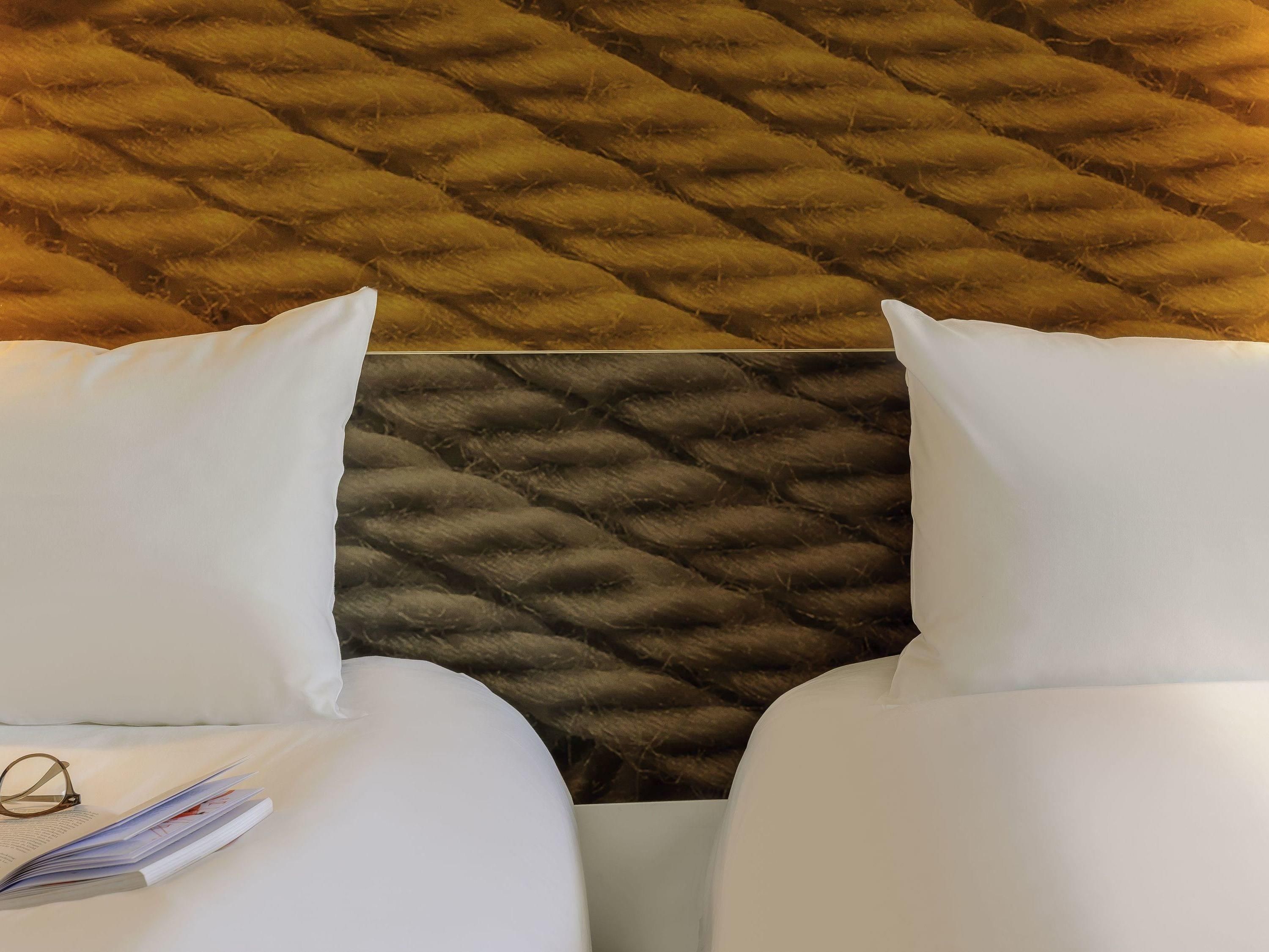 Une chambre d’hôtel avec une tête de lit capitonnée, deux oreillers blancs et une paire de lunettes posées sur un oreiller blanc.