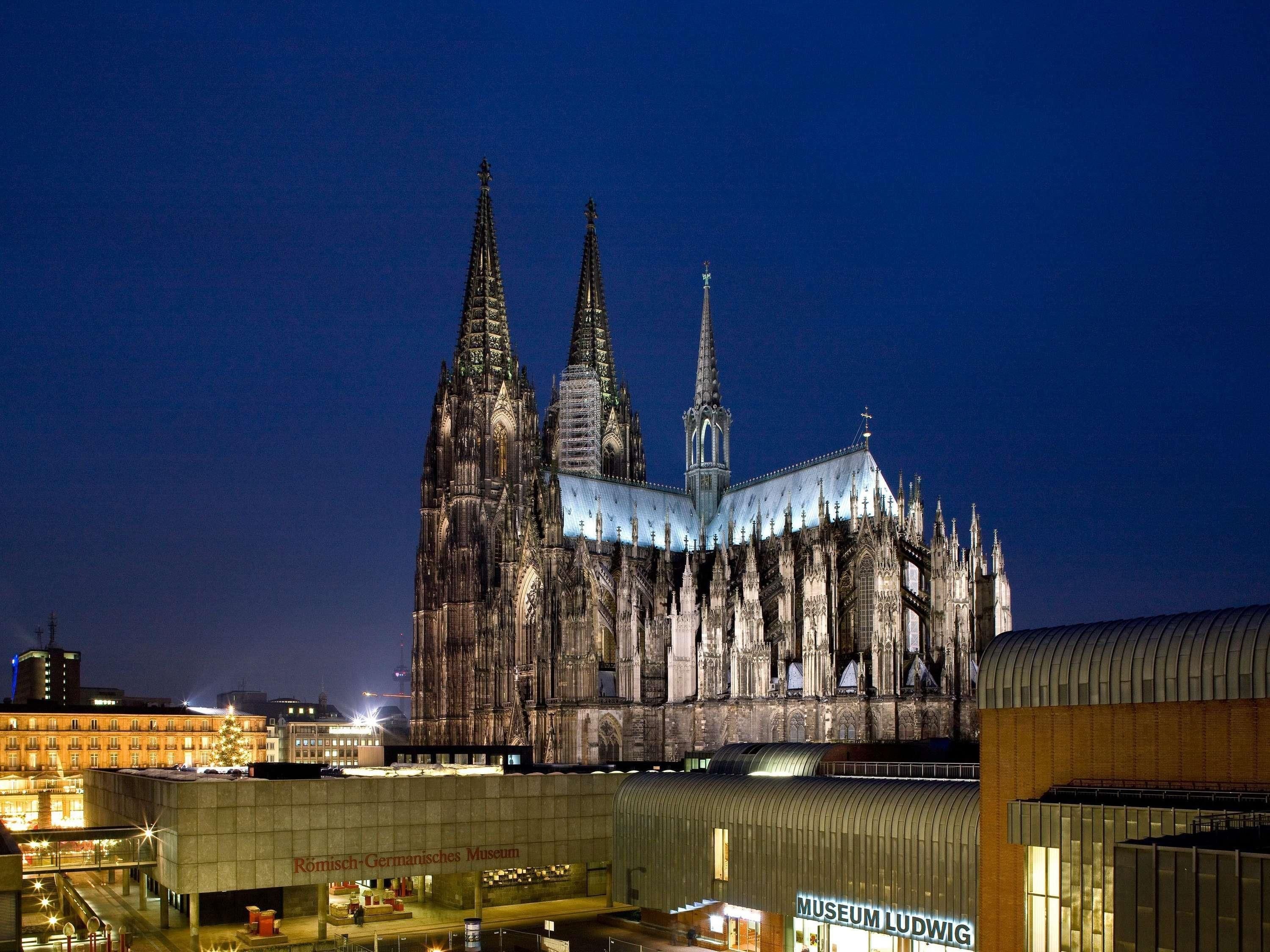 Une grande cathédrale dotée de deux flèches imposantes et d’une plus petite, le tout entouré de bâtiments modernes.