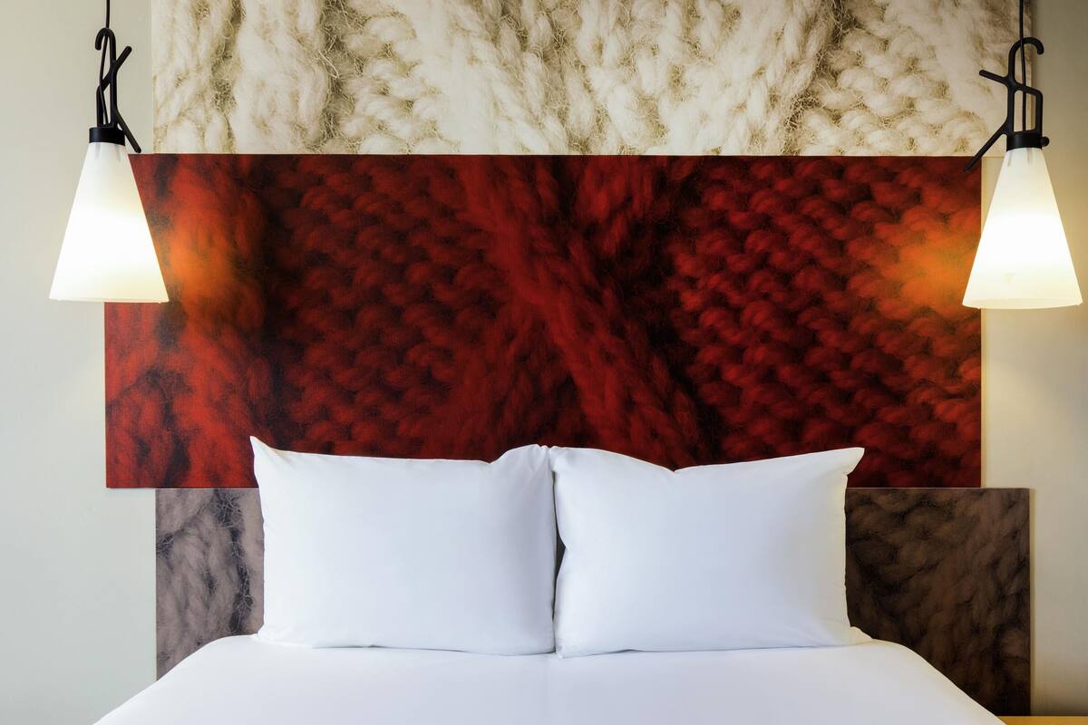 A bed with white linens, a red textured wall behind it, and two wall-mounted lamps.