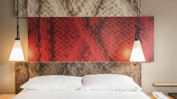 A hotel room with a bed, two pendant lights, a red and beige textured wall art, a white pillow, and a bedside table with a phone.