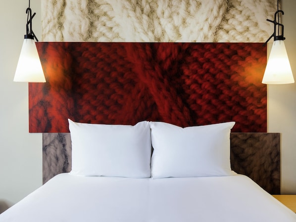 Un lit avec du linge de lit blanc, un mur rouge à texture derrière, et deux lampes fixées au mur.