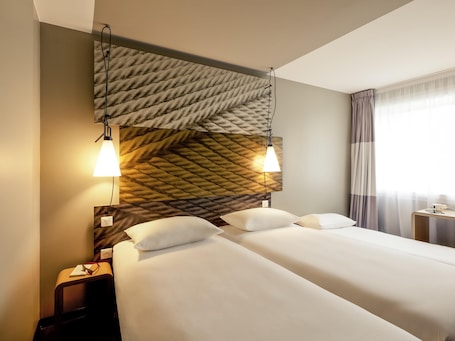 A hotel room with a large bed, two bedside lamps, a nightstand, and a window with curtains.