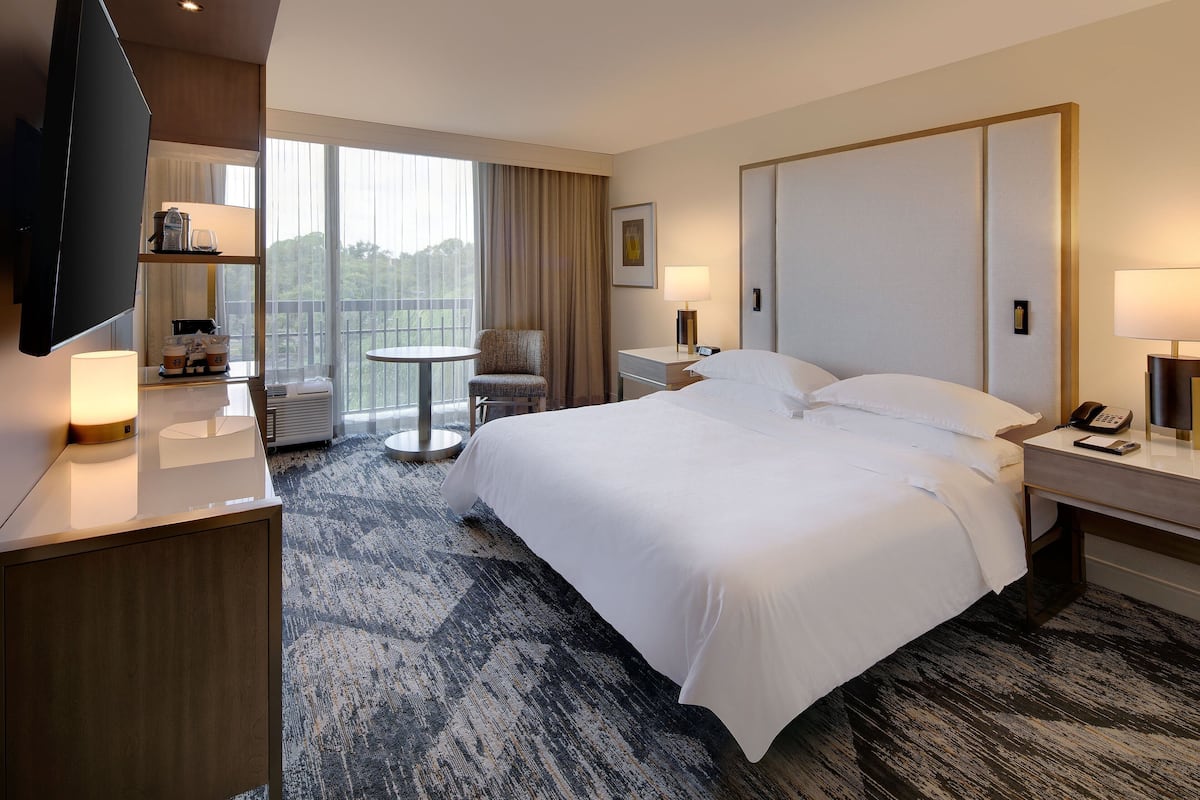 A hotel room with a large bed, a desk with a flat-screen TV, a chair, a small table, and a window with curtains.