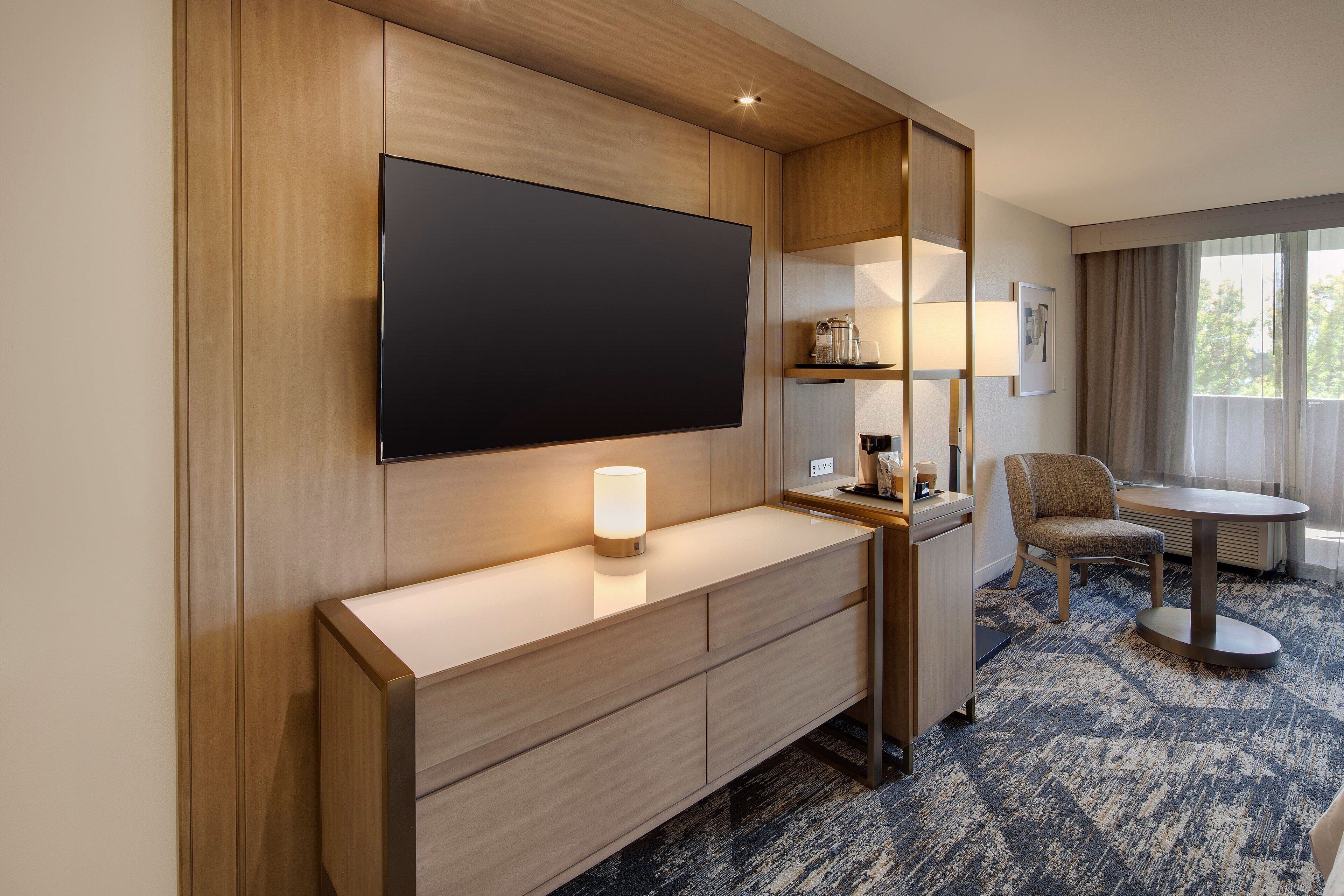 A hotel room with a large flat-screen TV mounted on the wall, a bedside table with a lamp, a chair, and a small round table.