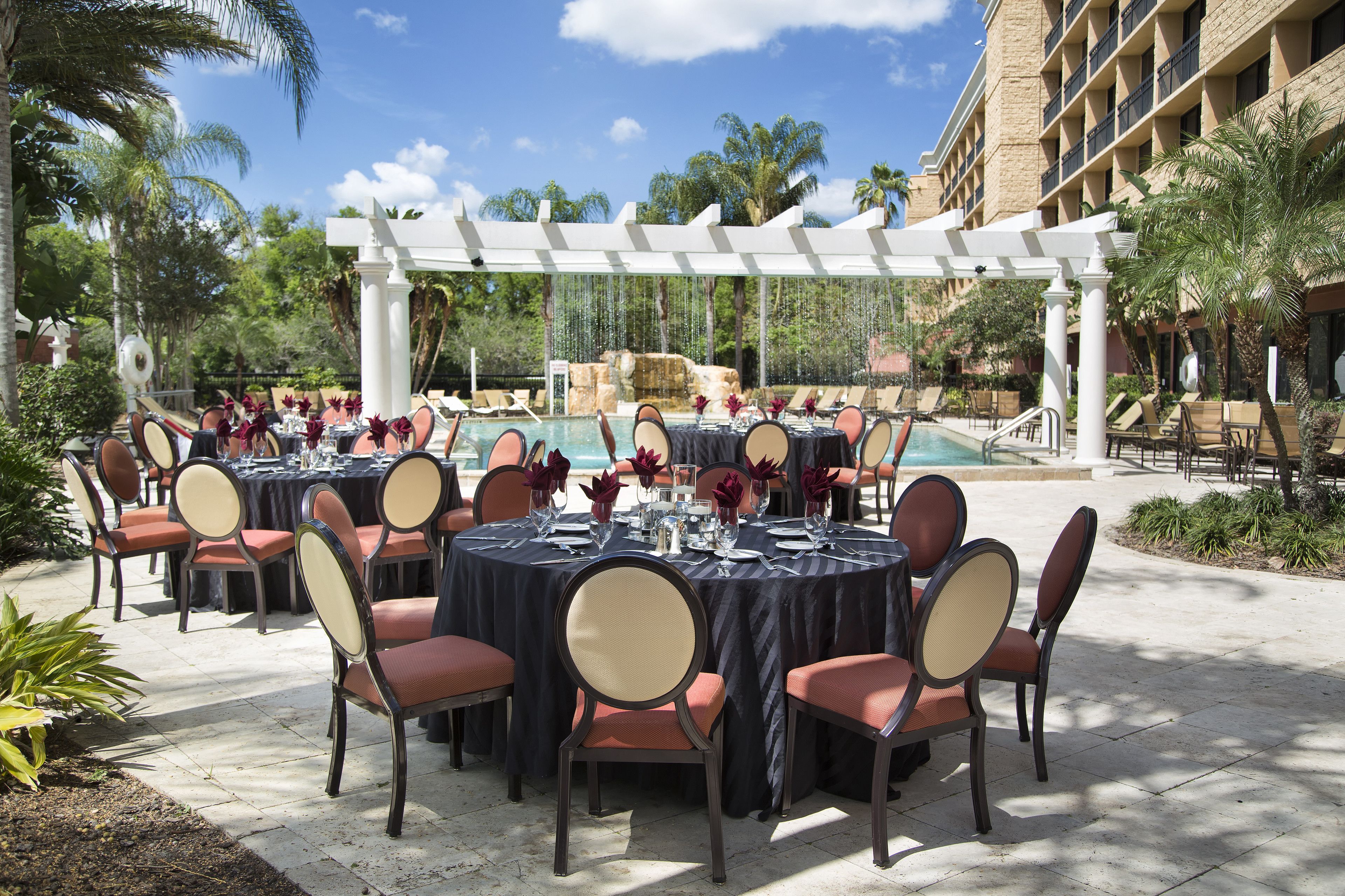 An outdoor dining area with round tables and chairs, a pool, and palm trees.