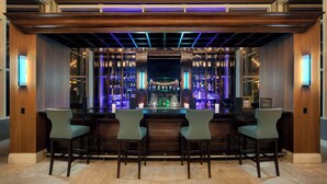 Una zona de bar con un mostrador de madera, dos taburetes y una estantería llena de botellas.