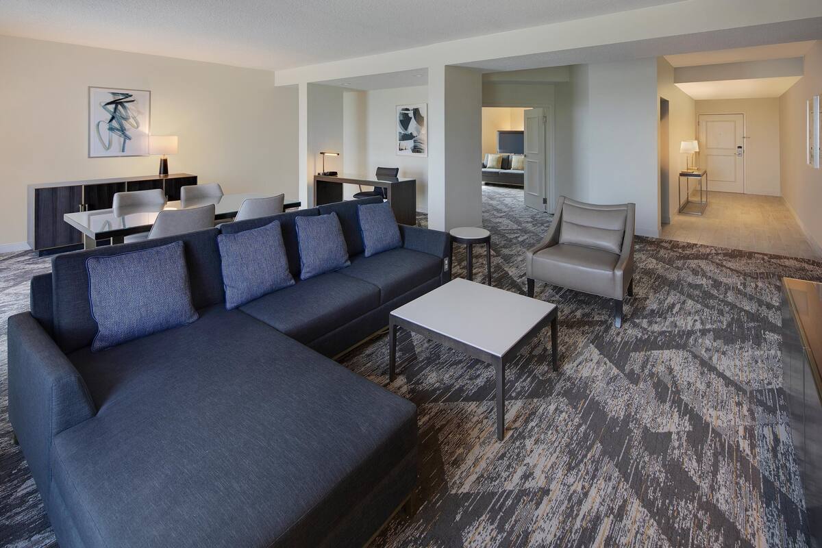 A modern hotel room with a sectional sofa, a small table, and a dining area with chairs and a desk.
