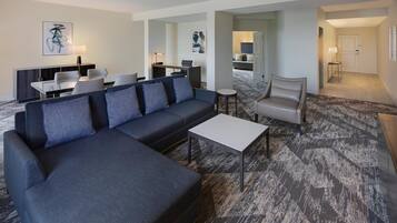A modern hotel room with a sectional sofa, a small table, and a dining area with chairs and a desk.