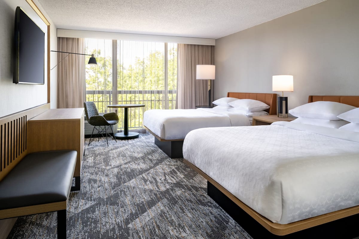 A hotel room with two beds, a desk, a chair, a TV, and a large window with curtains.
